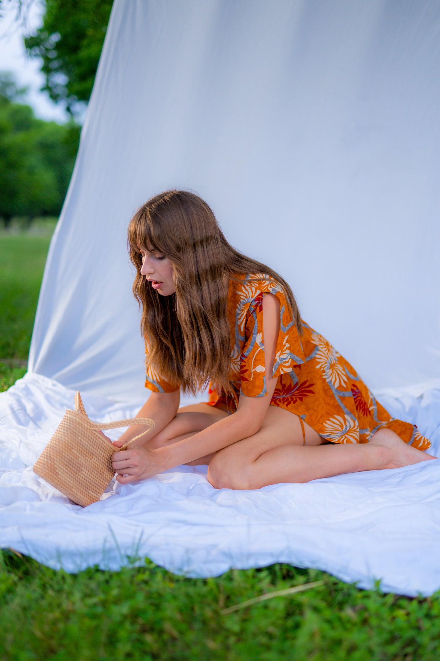 Woman in an orange dress sitting on a white blanket reading a book outdoors.
