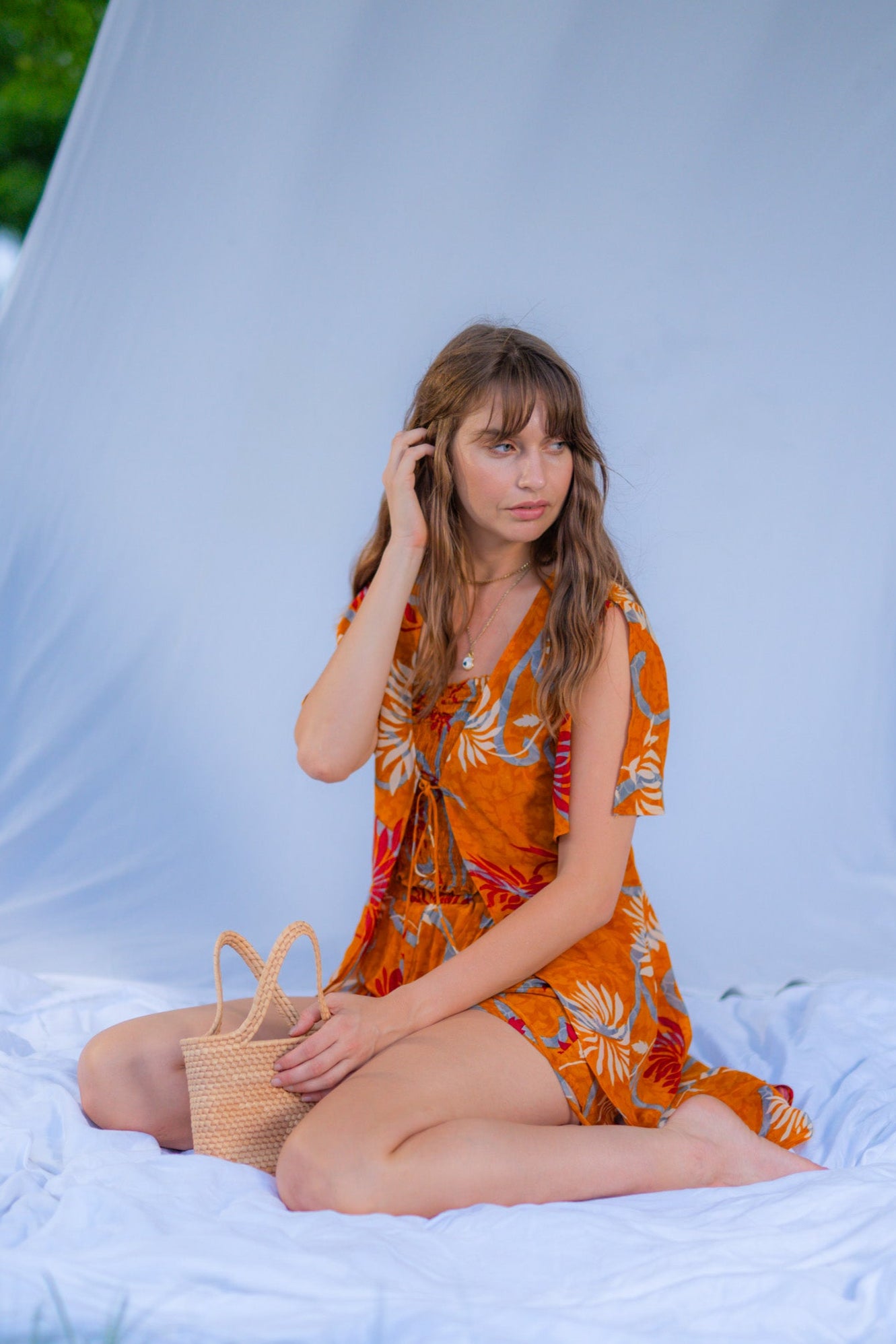 Woman in an orange floral dress sitting on a white blanket outdoors.
