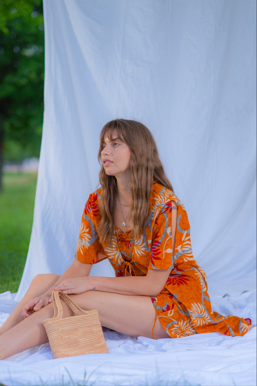 Woman in an orange dress sitting on a white blanket outdoors with a white sheet draped over trees.