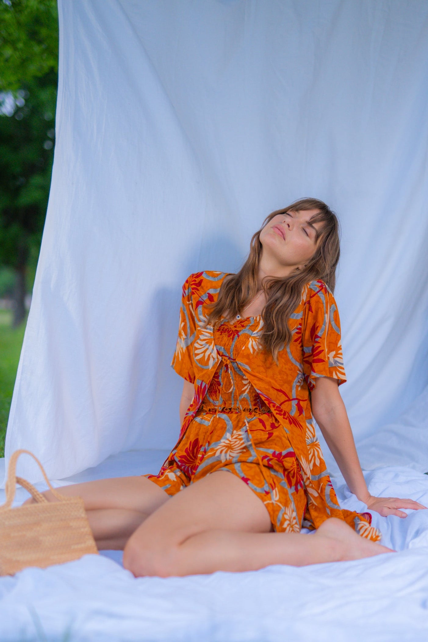 Woman in an orange dress sitting on a white blanket outdoors with a white curtain in the background.